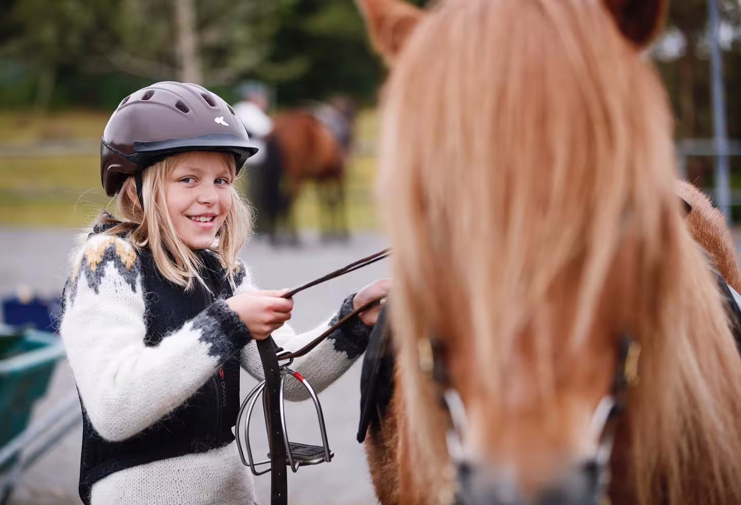 Smiling girl in riding helmet holding reins next to a chestnut horse at a stable