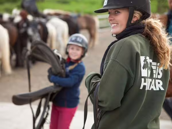 Smiling rider in green jacket and helmet holds reins at horseback riding lesson with child and horses in background