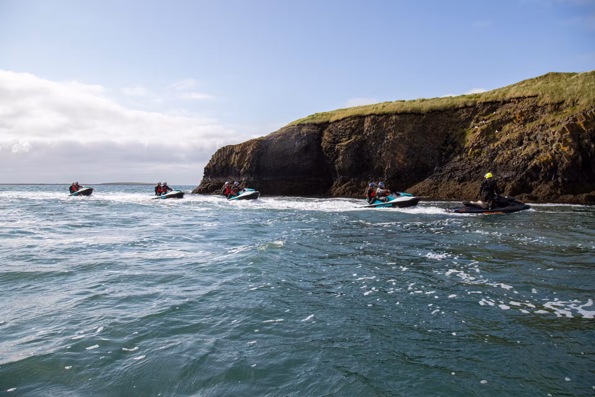 Group of jet skis riding along rocky Icelandic coastline with green cliffs on a clear day