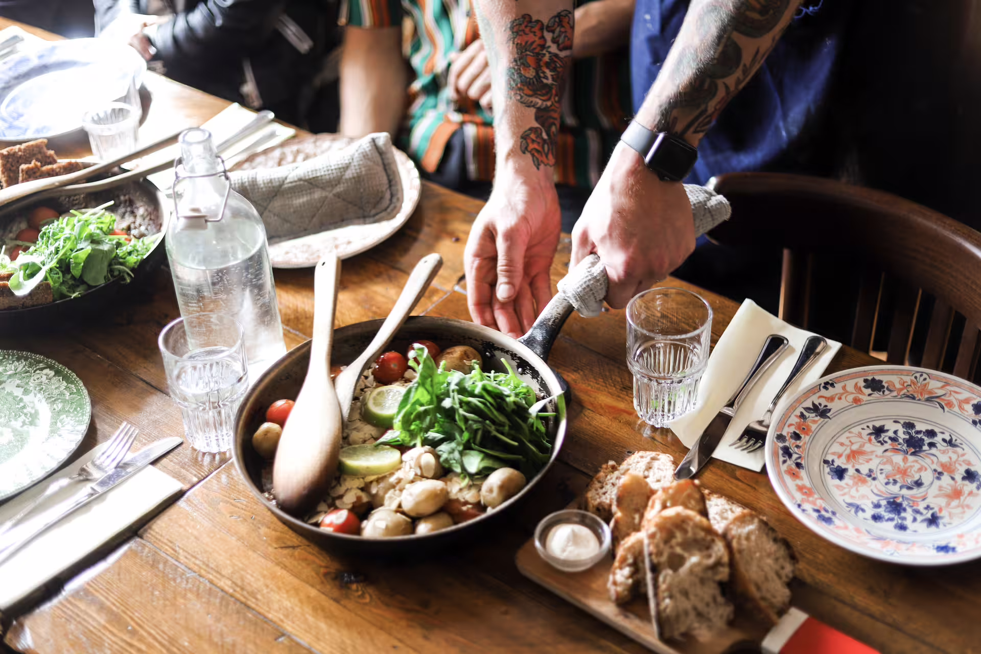 Tattooed server placing a skillet of fresh vegetables, tomatoes, and greens on a rustic wooden dining table