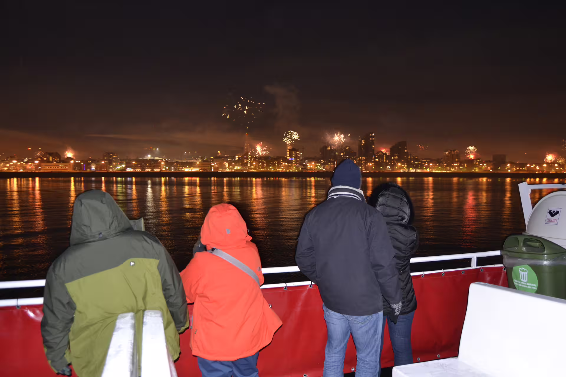 Passengers on a boat watching New Year's Eve fireworks over Reykjavik city skyline reflected in the sea