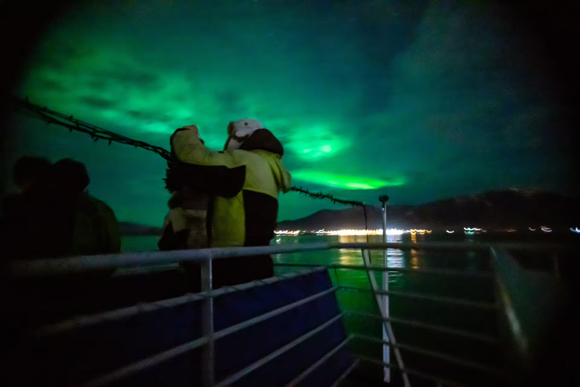 Couple embracing on boat deck watching vivid green northern lights over Reykjavik harbor at night