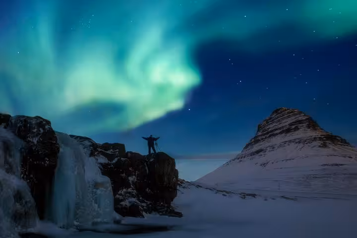 Person standing on snowy rocks with arms raised under vibrant green northern lights near Reykjavik, Iceland