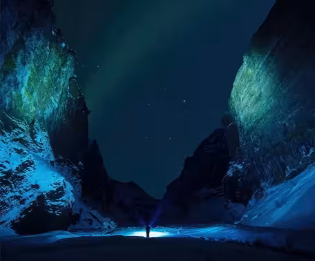 Solo traveler with flashlight standing between snowy canyon walls under northern lights in Iceland
