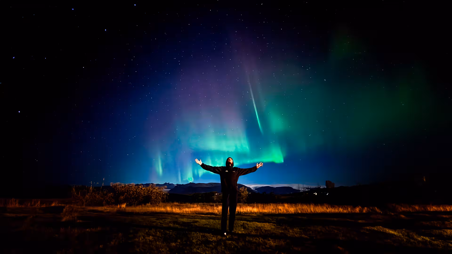 Person standing with arms outstretched under vivid green and purple northern lights in Reykjavik night sky