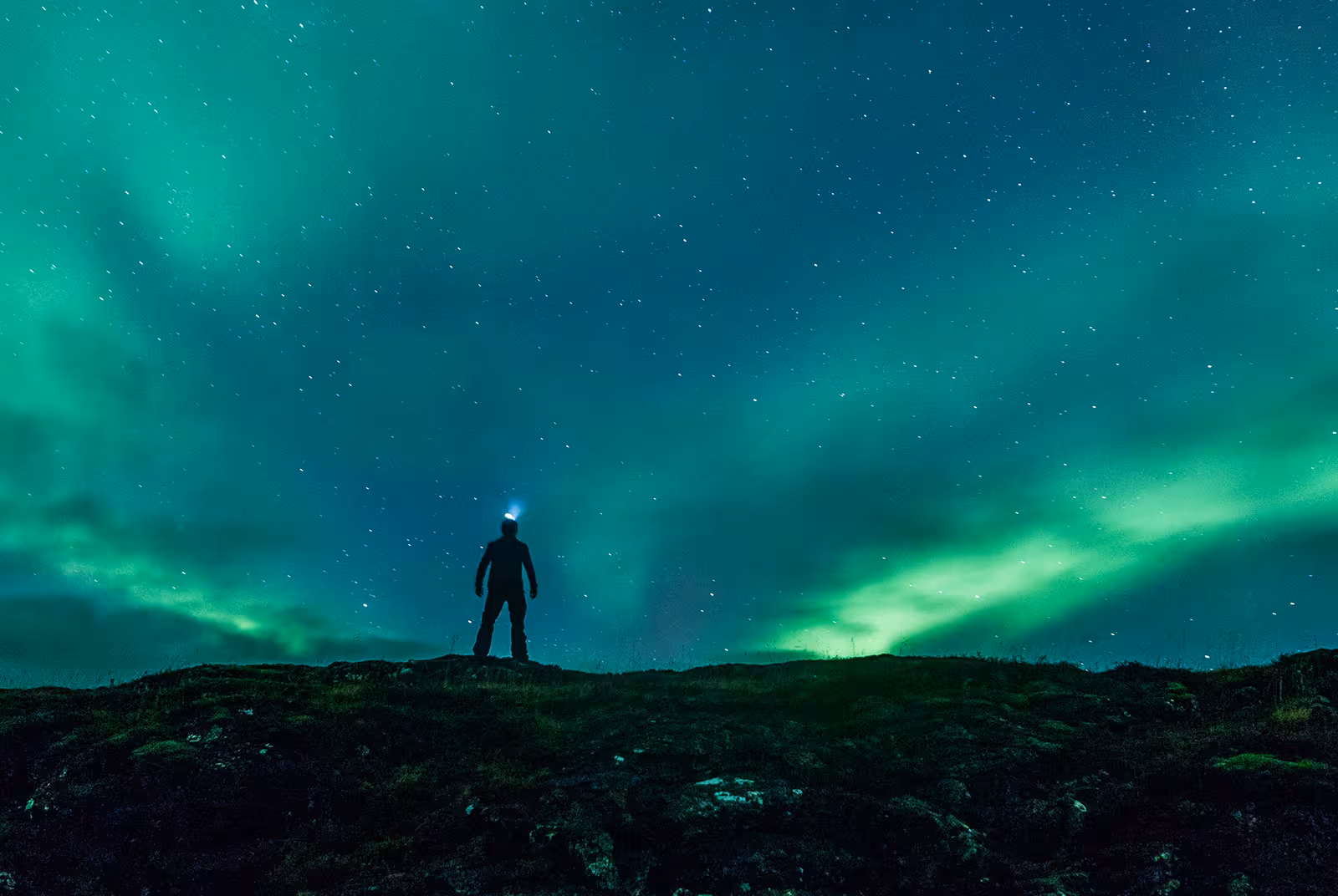 Silhouetted person with headlamp standing on hill beneath vivid green northern lights in Reykjavik night sky