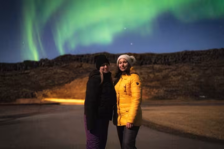 Two women smiling under vivid green northern lights in Reykjavik night sky