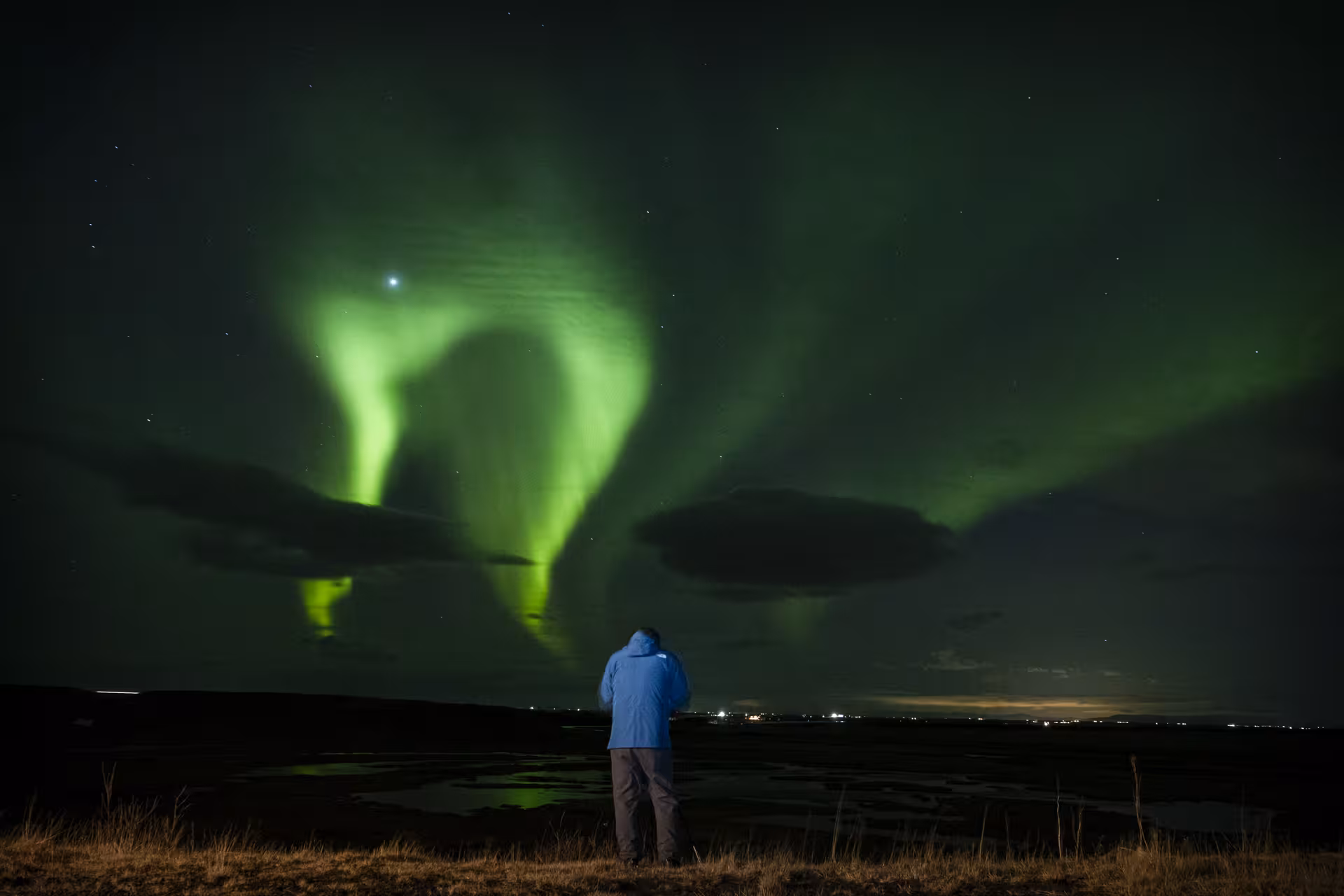Person in blue jacket watching vivid green northern lights aurora borealis over Reykjavik night sky