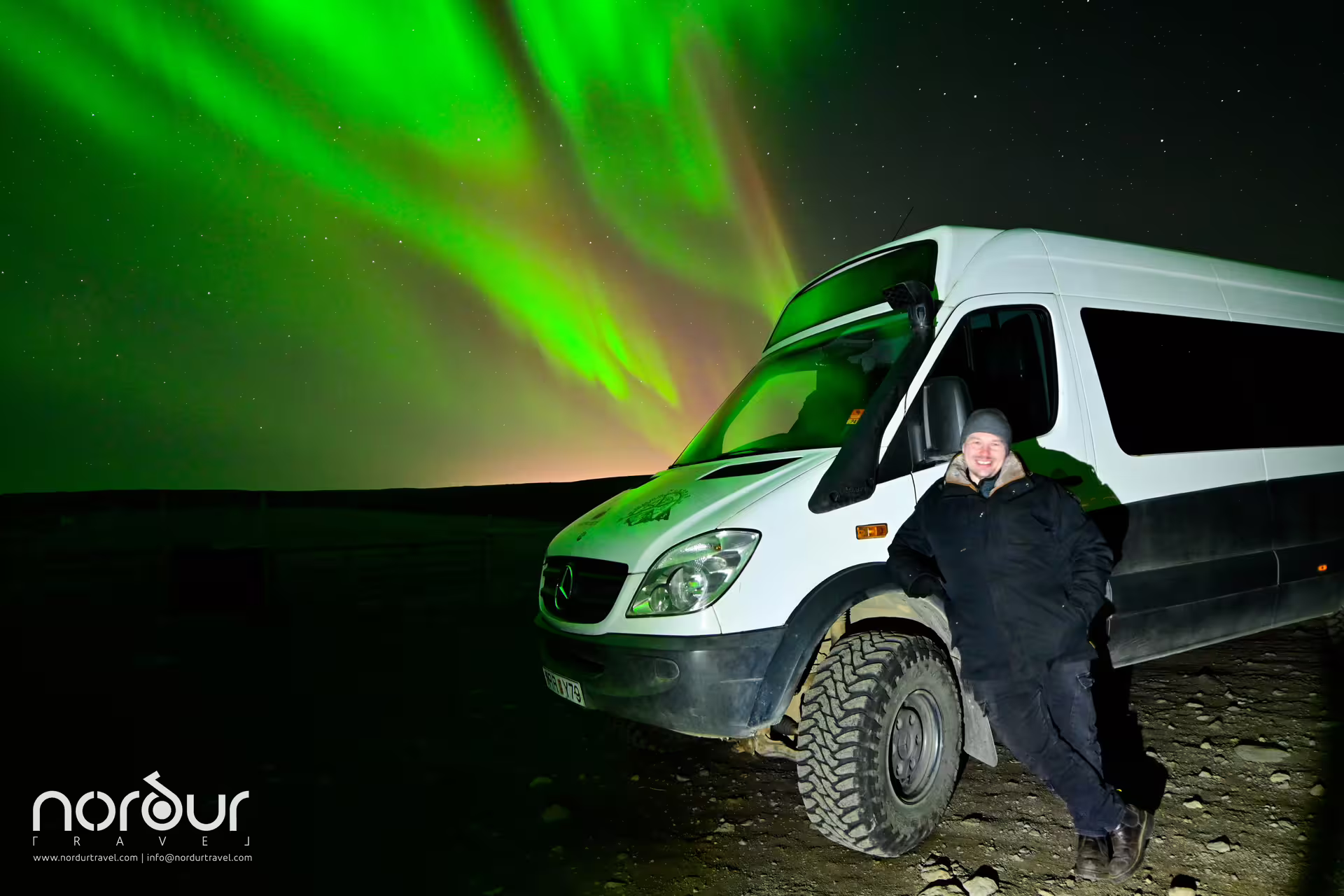 Smiling guide stands beside a white tour van under vivid green aurora borealis on a Reykjavik northern lights tour