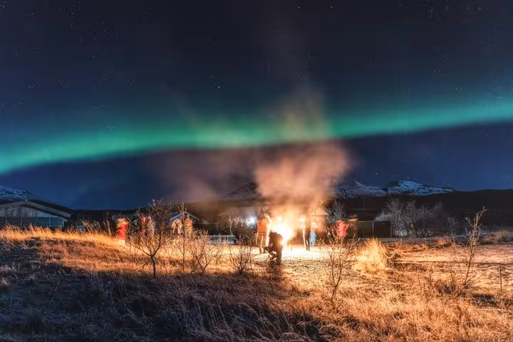 Group gathered around bonfire under glowing green northern lights in Reykjavik night sky