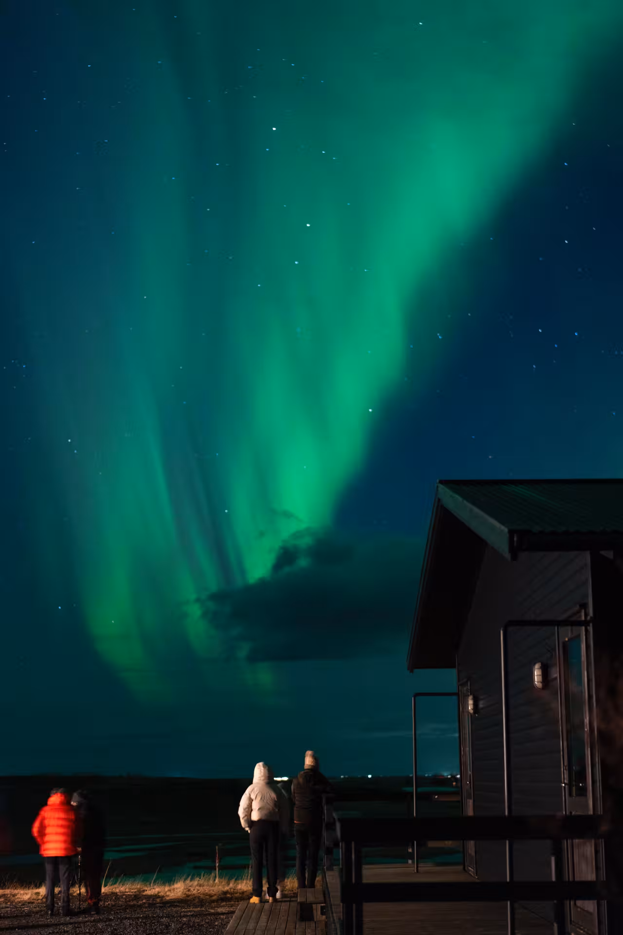 Three people in winter jackets watching vibrant green northern lights from a cabin deck in Reykjavik