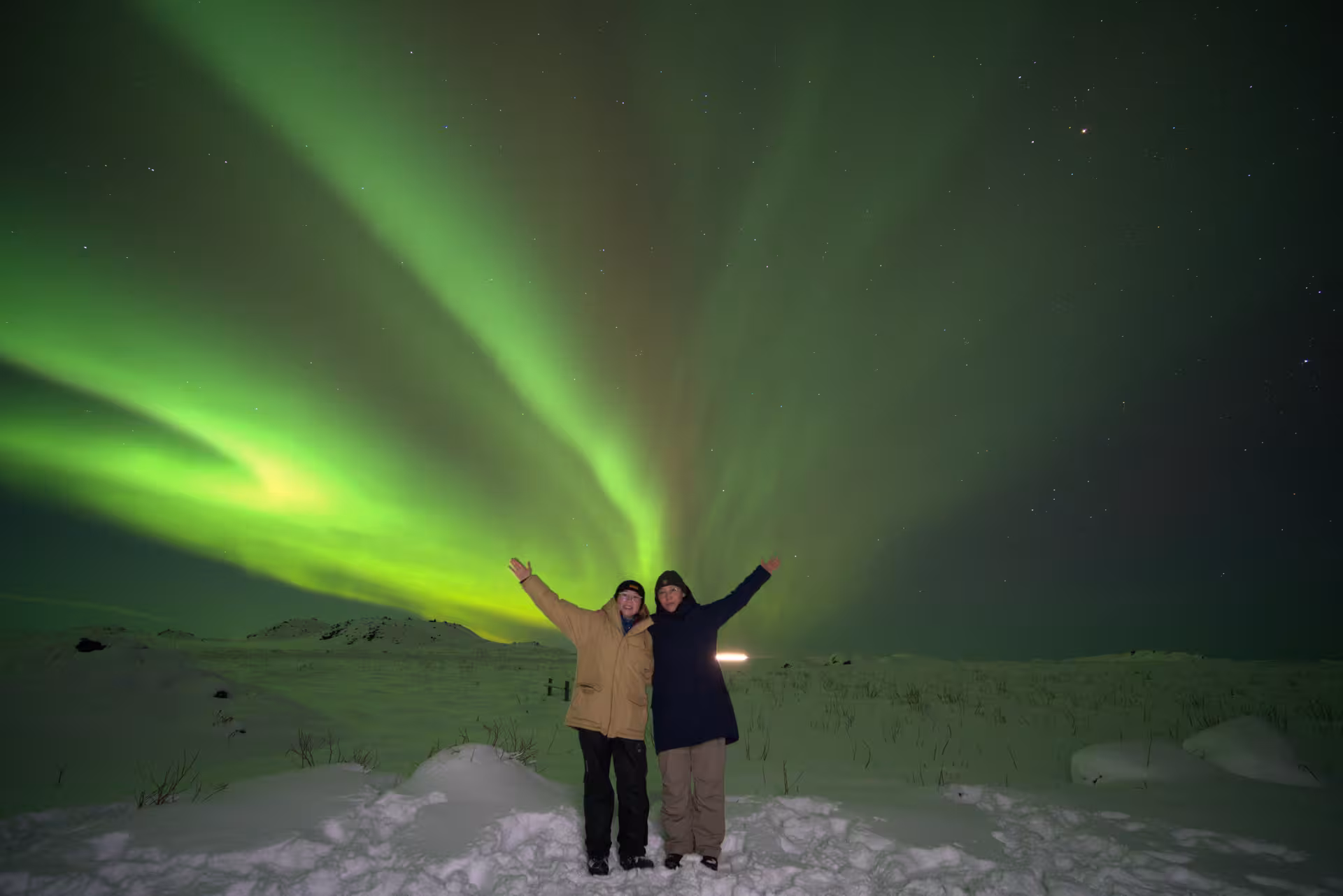 Two tourists with arms raised under vibrant green northern lights over snowy Iceland landscape at night