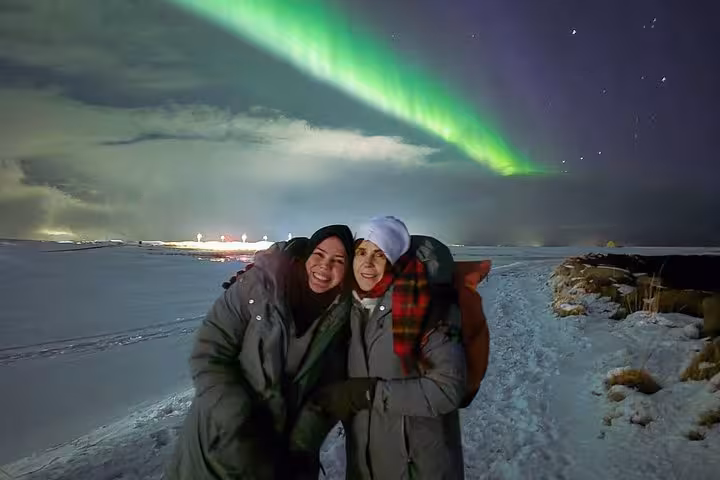 Two smiling women posing under vibrant green northern lights in snowy Reykjavik night landscape