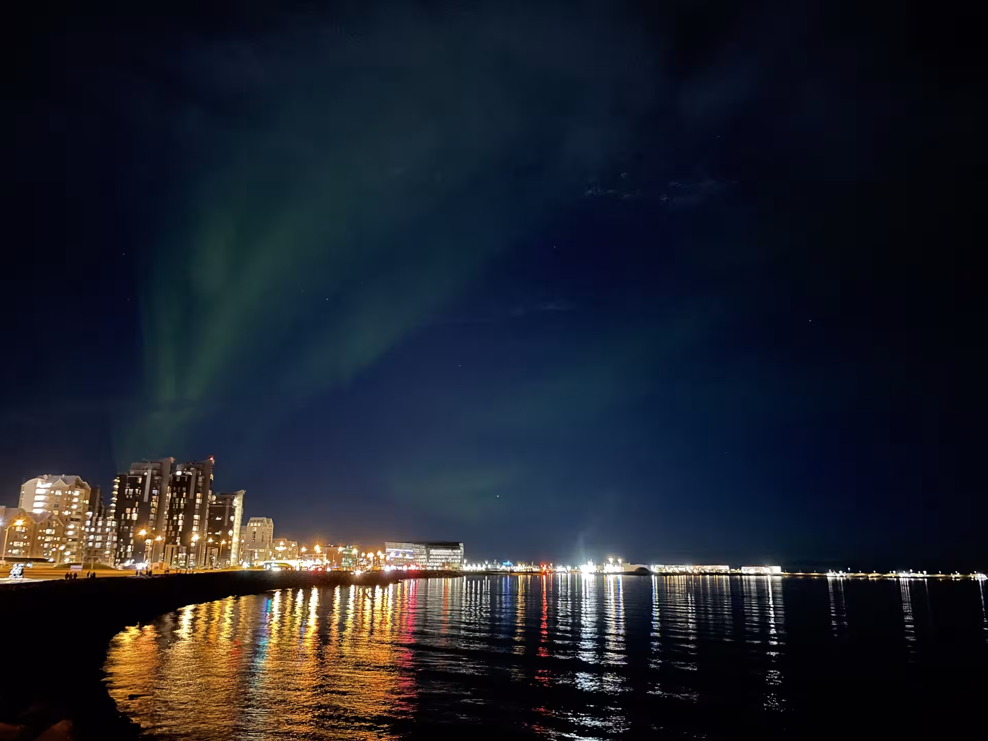 Northern lights glowing green over Reykjavik waterfront skyline at night with colorful city reflections