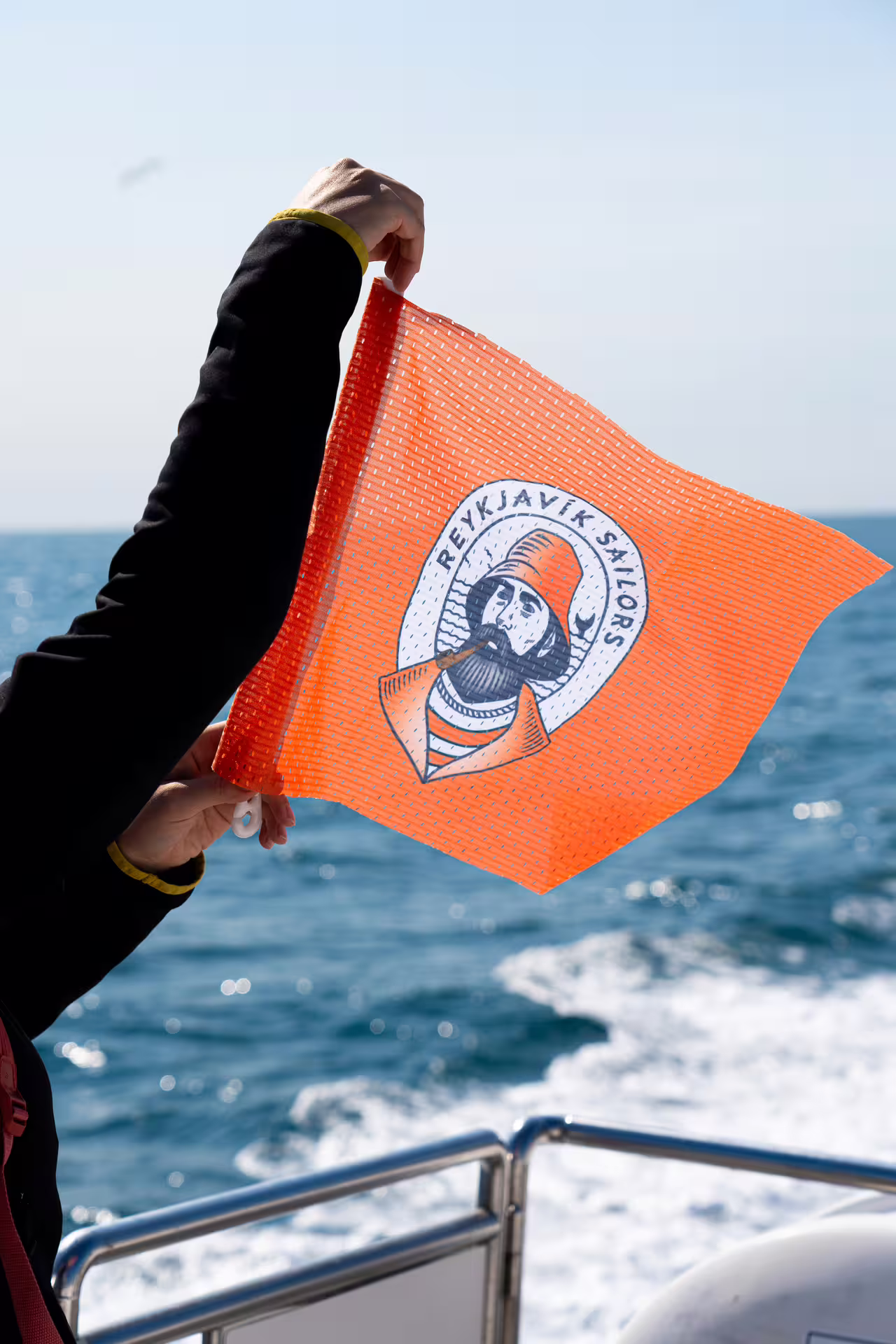 Person holding orange Reykjavik Sailors flag on boat with ocean waves in background