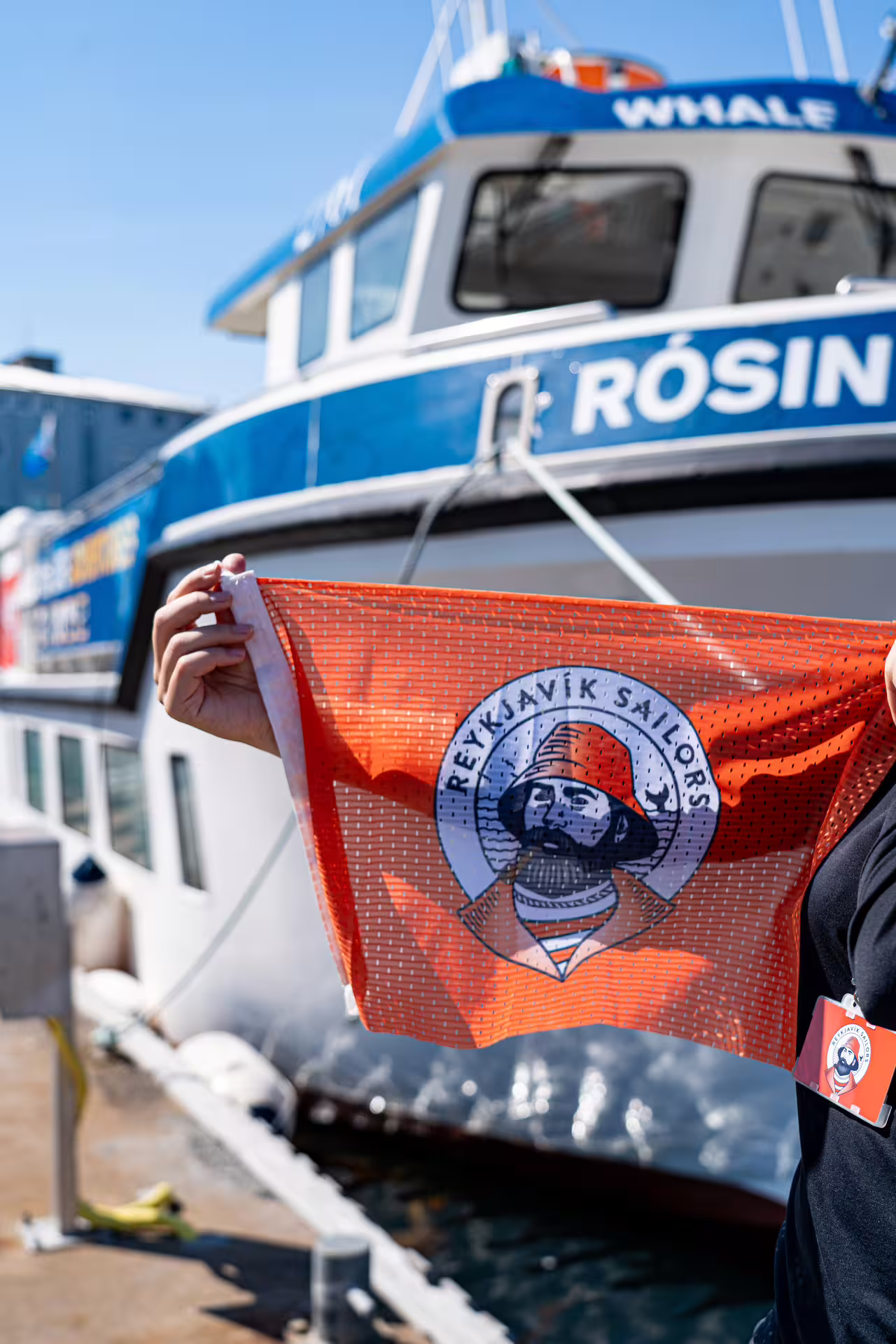 Person holding orange Reykjavik Sailors flag at Old Harbour with whale watching boat Rósin in background