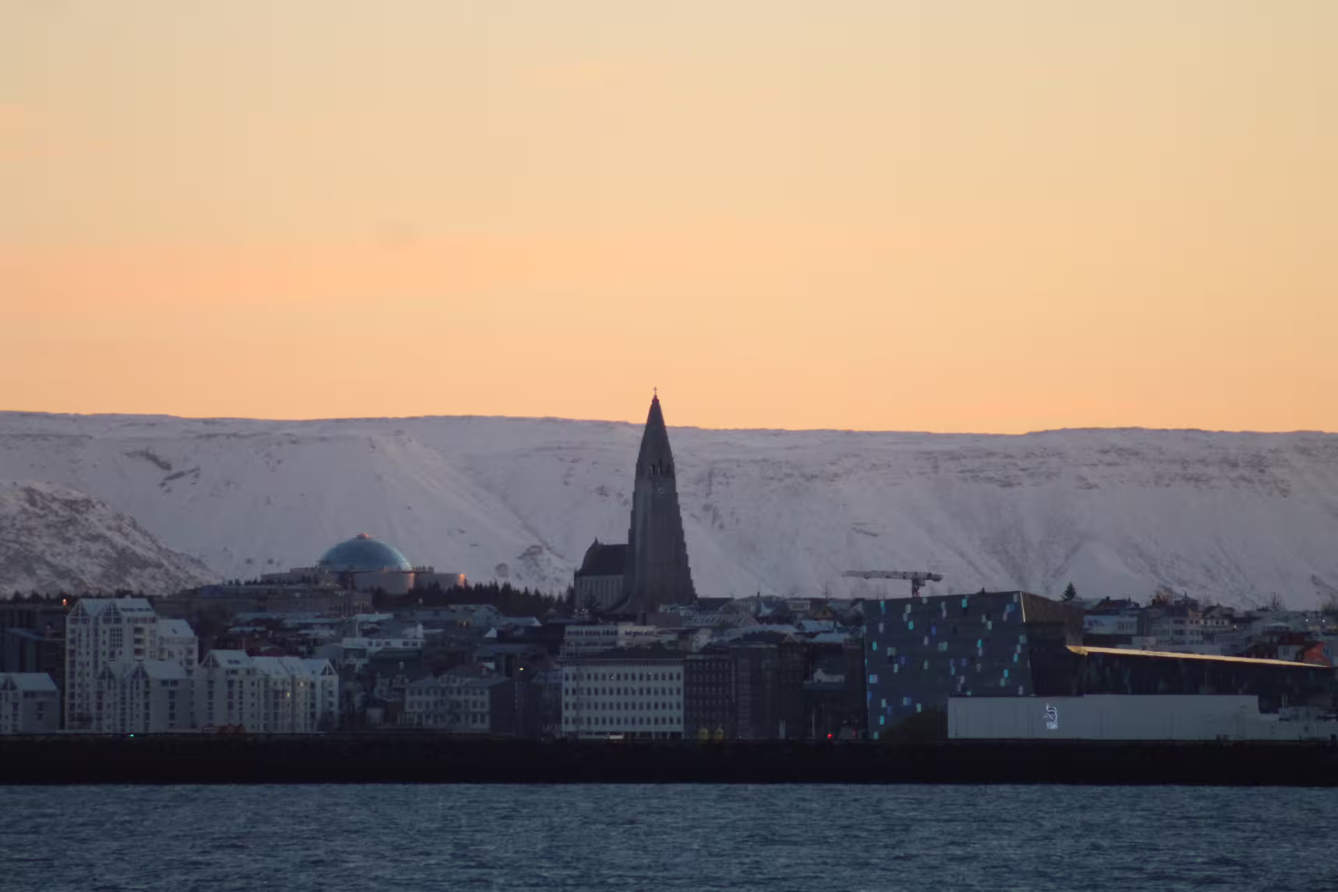 Reykjavik cityscape at sunset with Hallgrímskirkja church, Harpa concert hall, and snow-covered mountains across the harbor