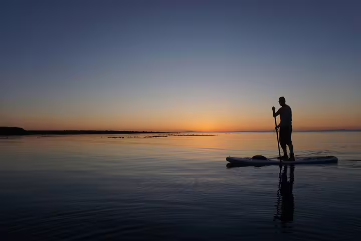 Silhouetted paddle boarder gliding on calm waters during a vibrant Reykjavik sunset, with a lighthouse in the background.