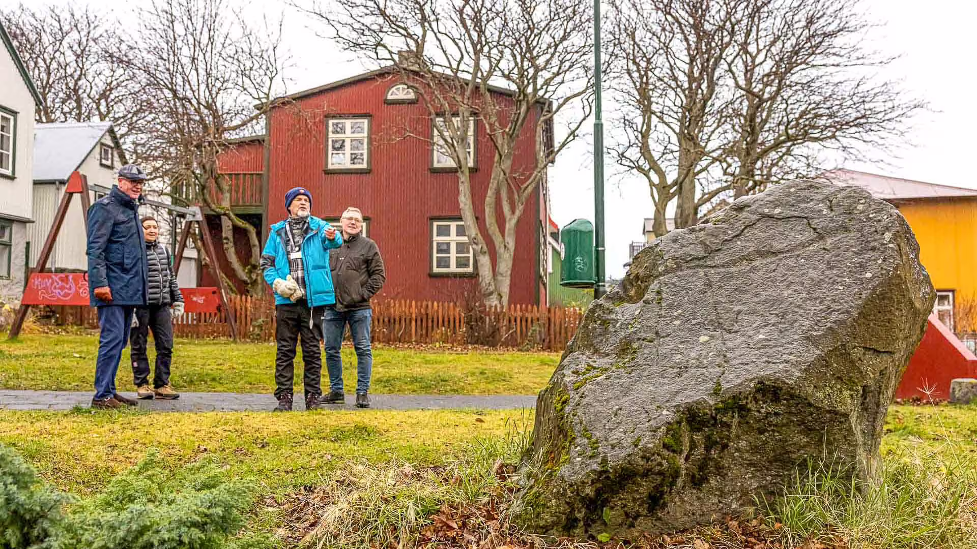 Tour guide pointing out landmarks to visitors near a large boulder in a Reykjavik park with red building