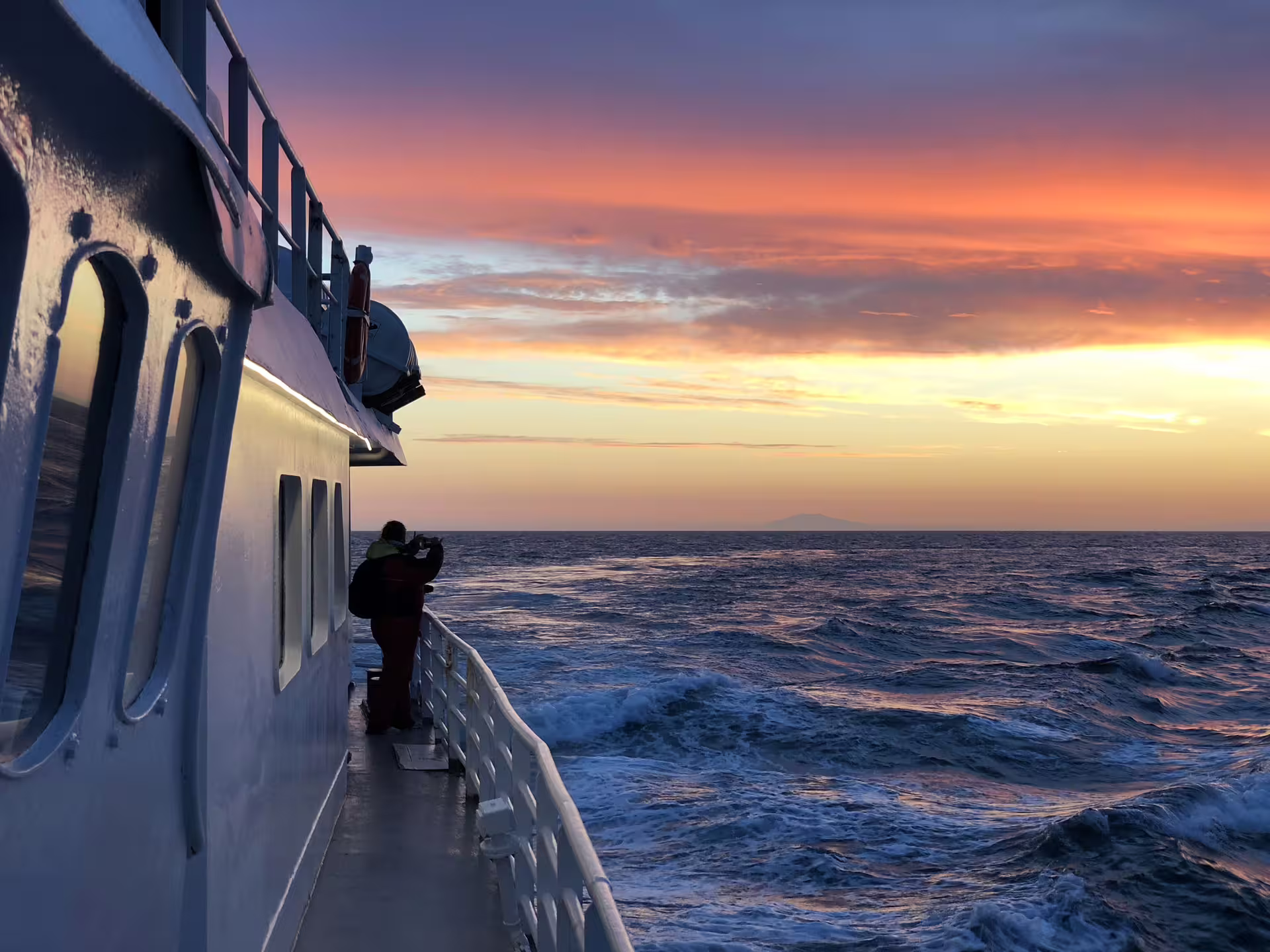 Photographer on whale watching boat deck capturing dramatic midnight sun sunset over rough ocean in Reykjavik