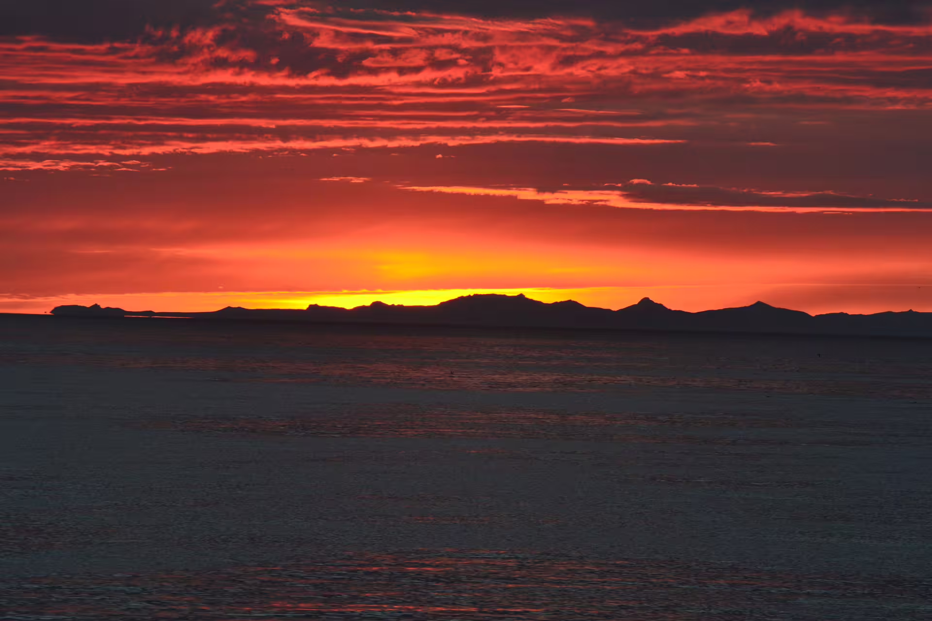 Fiery red and orange midnight sun sunset over mountain silhouettes reflected on calm ocean waters in Reykjavik