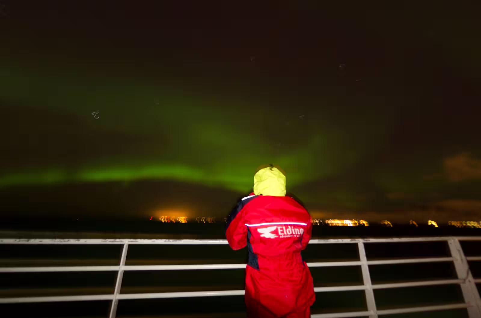 Guest in red Eldino jacket watching green northern lights from whale watching boat deck in Reykjavik