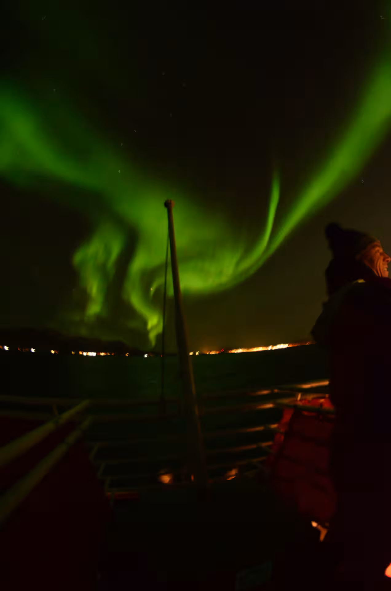 Person on boat deck watching vibrant green northern lights dancing over Reykjavik waters at night