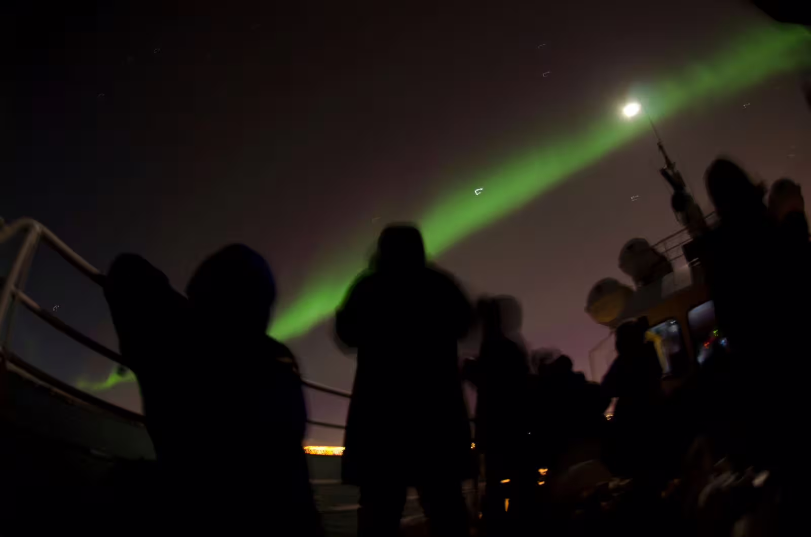 Silhouettes of whale watching tour guests on boat deck under green northern lights and moon in Reykjavik