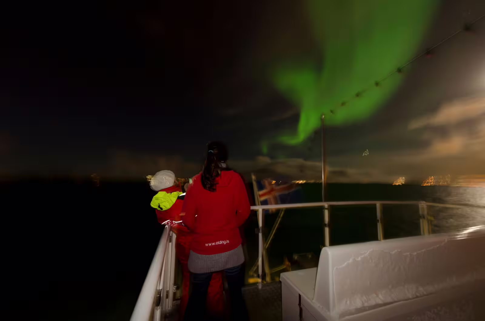 Guide in red Elding jacket watching vivid green northern lights from whale watching boat deck in Reykjavik
