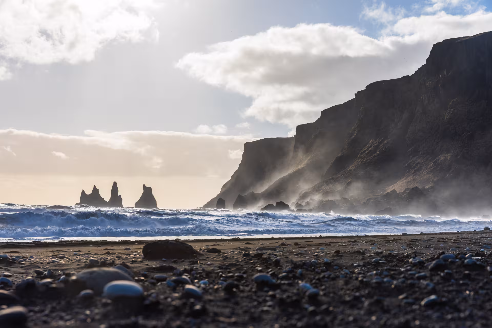 Sunlit Reynisdrangar sea stacks and misty cliffs along the black sand beach of Iceland's South Coast.