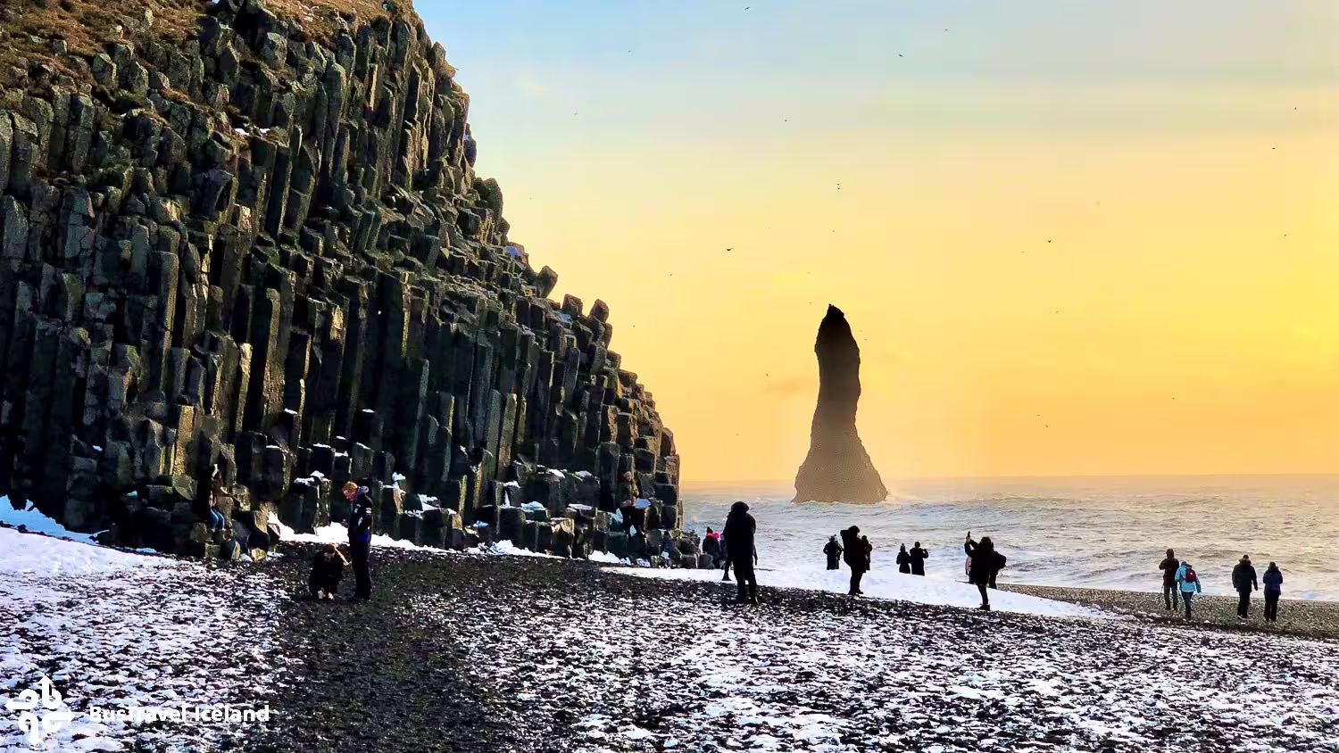 Tourists on Reynisfjara black sand beach with basalt columns and sea stack at golden sunset, Iceland