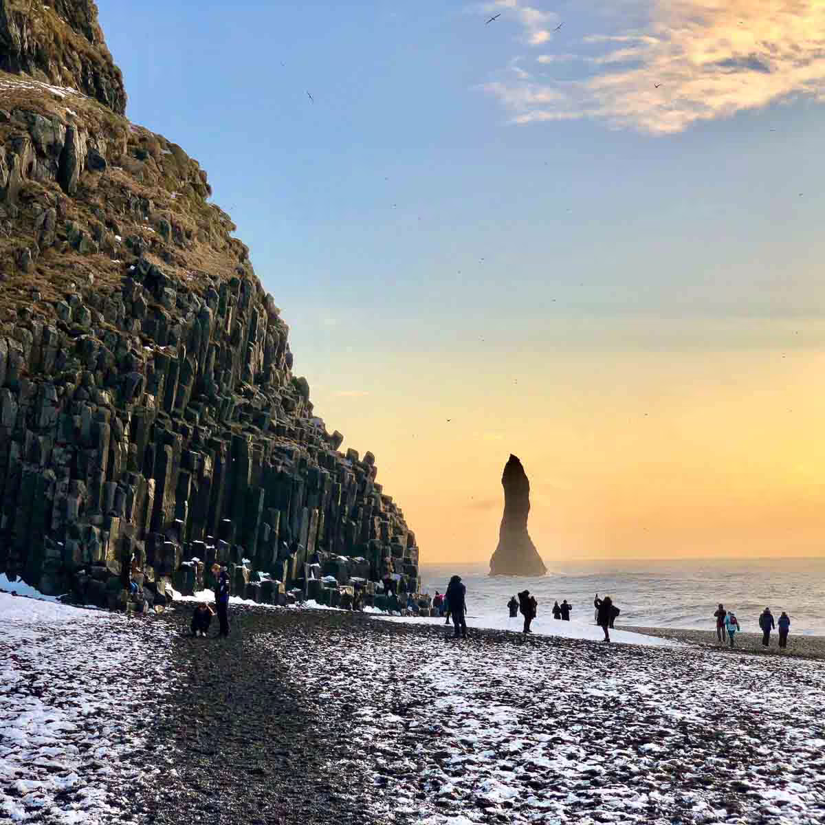 Tourists walking on Reynisfjara black sand beach with basalt columns and sea stack at sunset, Iceland