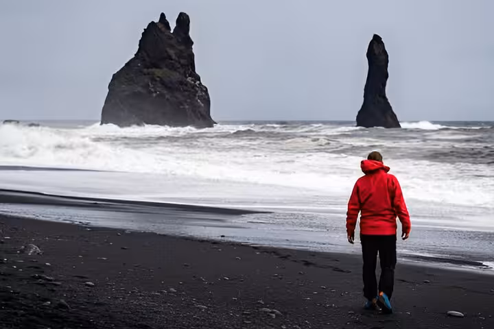 Person in red jacket walking on Reynisfjara black sand beach with volcanic rock stacks and waves, South Coast Iceland