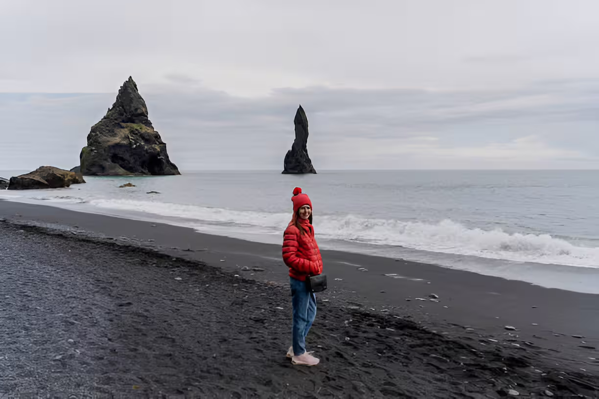 Woman in red jacket on Reynisfjara black sand beach with volcanic rock stacks, Iceland South Coast