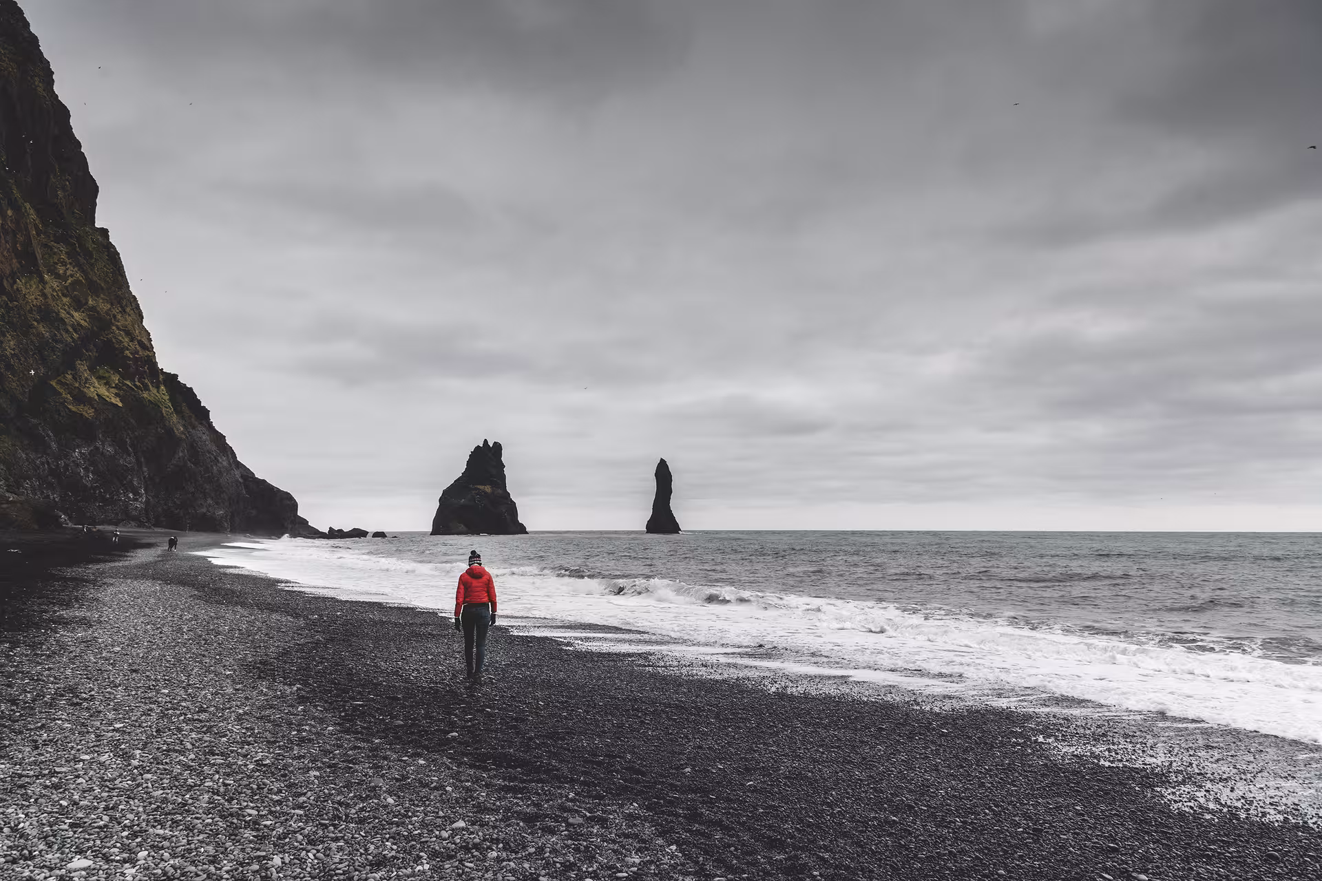 Person in red jacket walking on Reynisfjara black sand beach with sea stacks and dramatic cloudy sky, Iceland