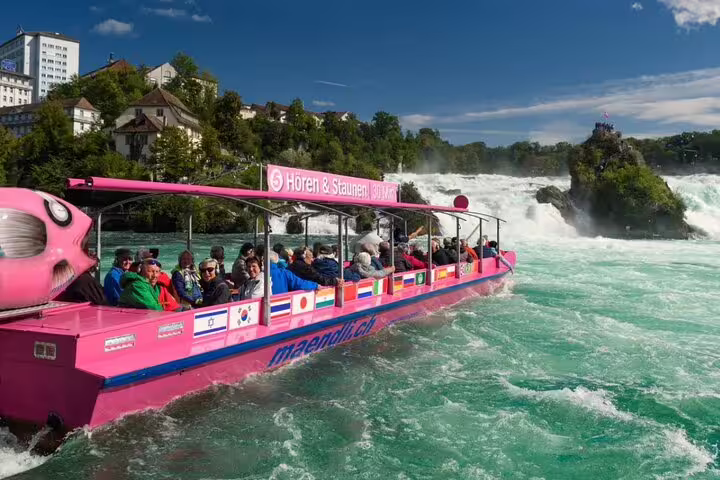 Tourists enjoy a scenic boat ride near the majestic Rhine Falls, surrounded by lush greenery under a clear blue sky.