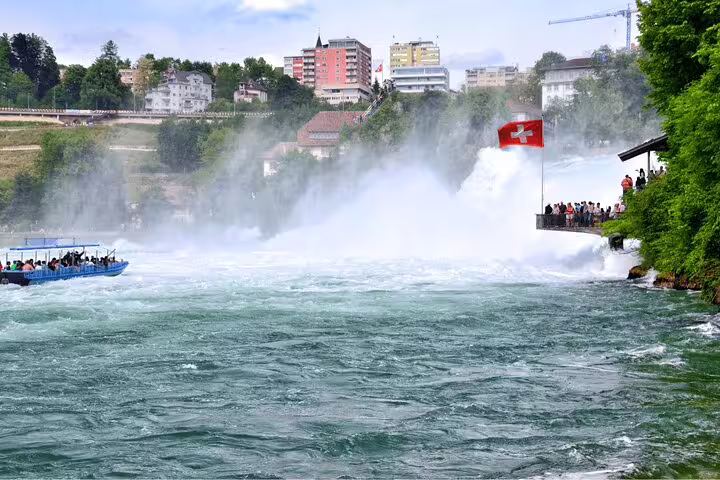 Tourists enjoy scenic views of Rhine Falls from the river with Swiss flag and lush surroundings on Zurich day tour.
