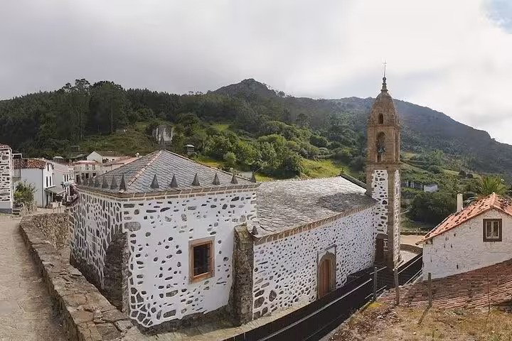 Scenic view of a charming stone church in Rías Altas surrounded by lush greenery and mountainous backdrop.