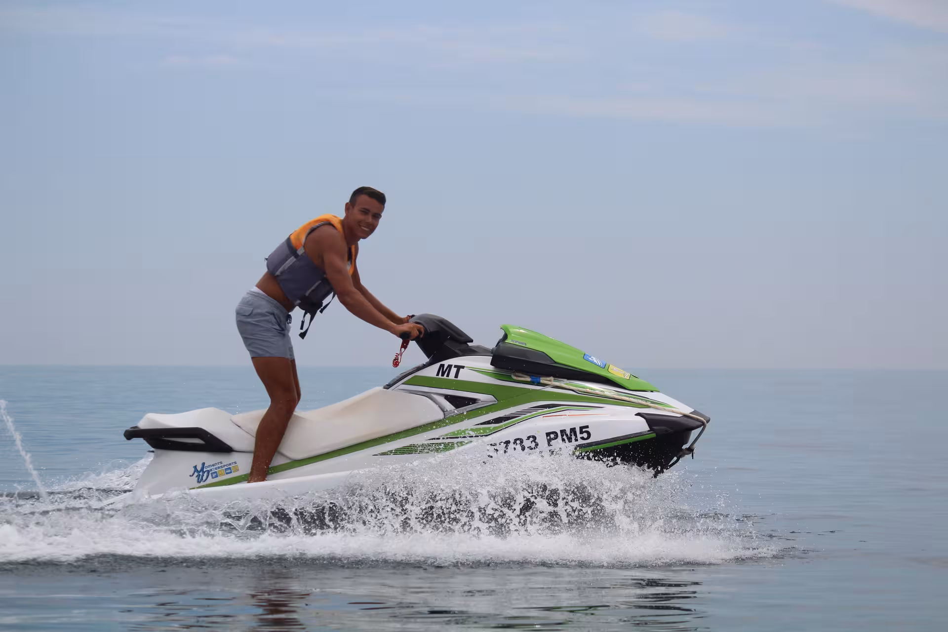 Excited rider wearing a life jacket standing on a green and white jet ski over ocean waves.