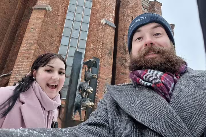Two smiling tourists taking a selfie in front of a historic brick church with bronze animal sculptures in Riga