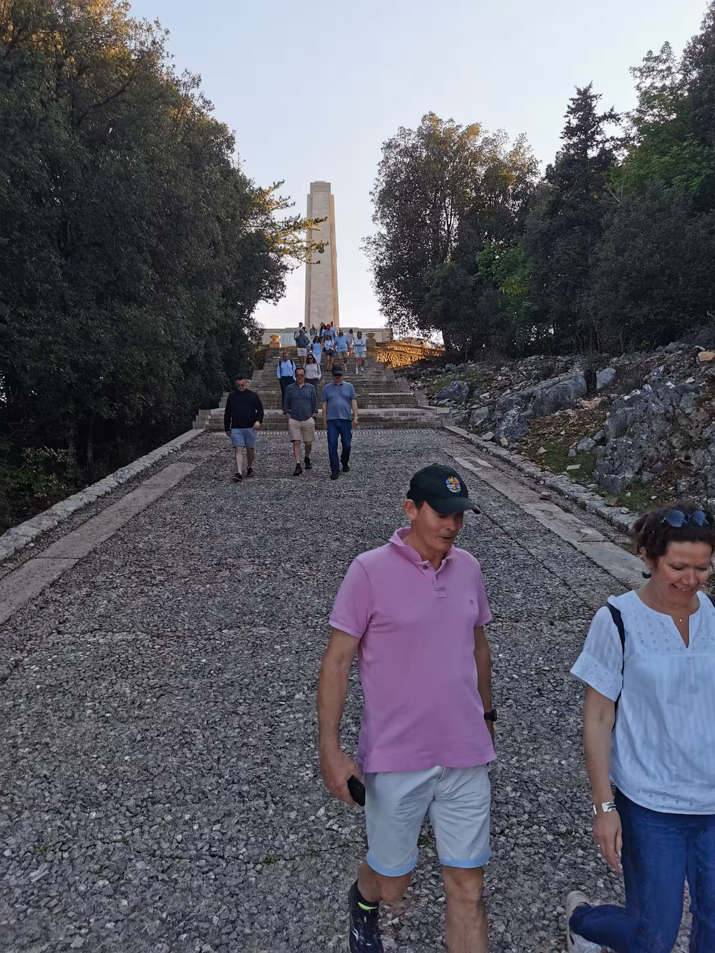 Tourists descend a rocky path surrounded by trees on L'Anello della Collina Imprendibile, with a monument in the distance.