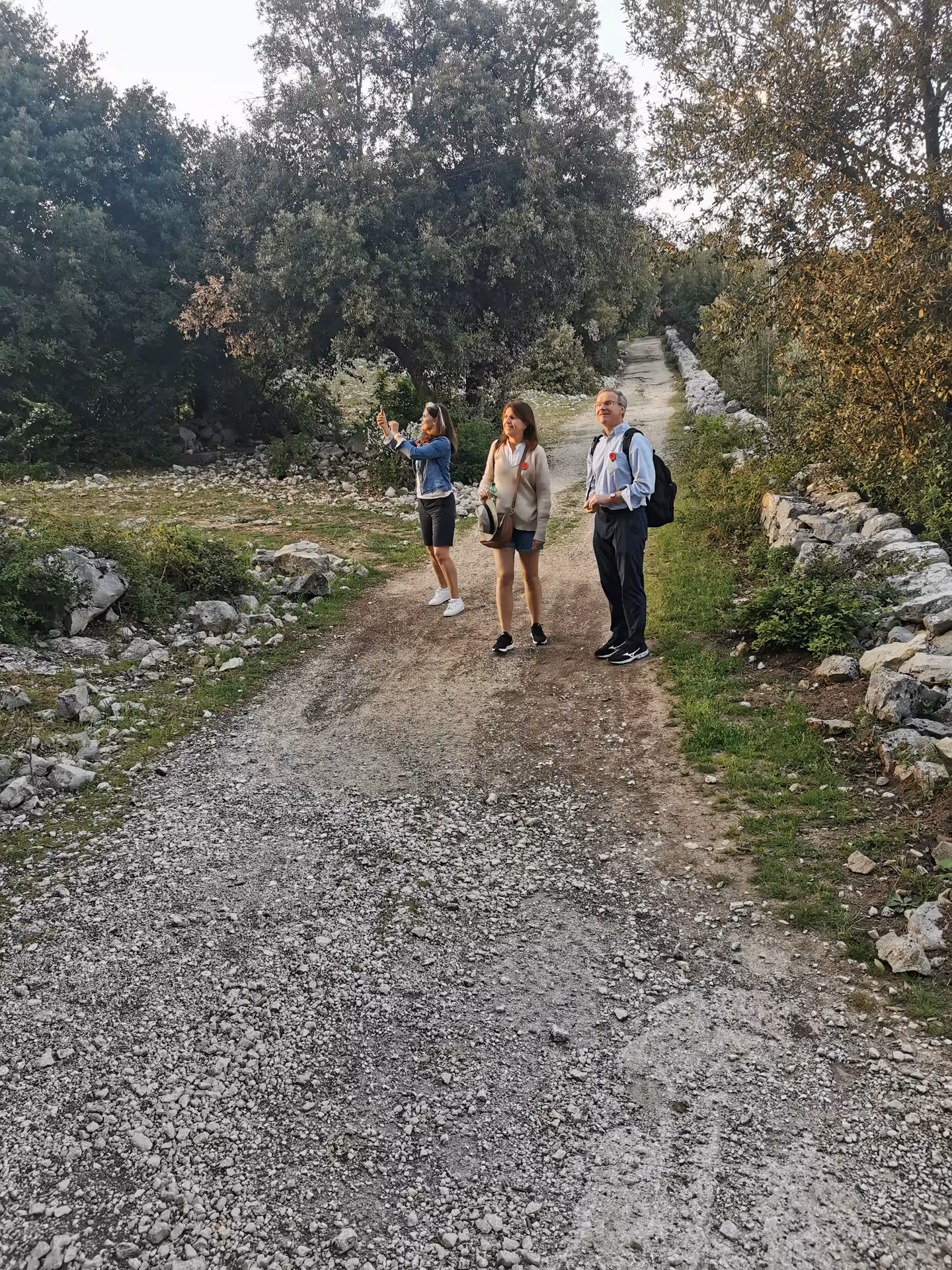 Tourists exploring a rustic path in L'Anello della Collina Imprendibile, capturing nature's beauty with their phones.
