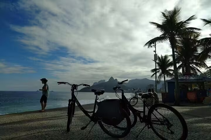 Scenic bike ride along Rio's coastline with ocean view, palm trees, and distant mountains on a cloudy day.