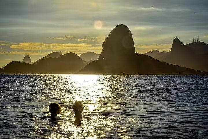 Silhouetted swimmers in Rio's ocean with stunning sunset views of Sugarloaf Mountain, ideal for romantic boat tours.