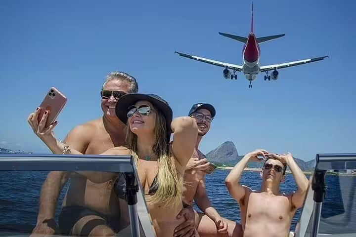 Group enjoying a Rio boat tour with barbecue, capturing a selfie as a plane soars above in sunny weather.