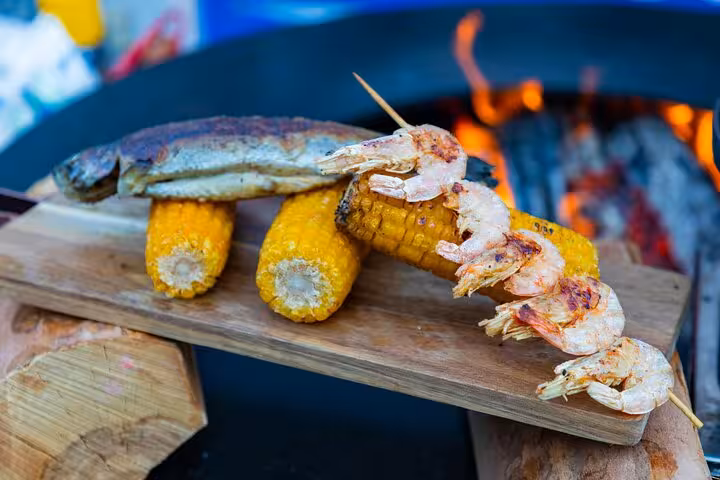 Grilled shrimp, fish, and corn on a wooden platter by a fire pit, part of a Rio seafood cooking class experience.