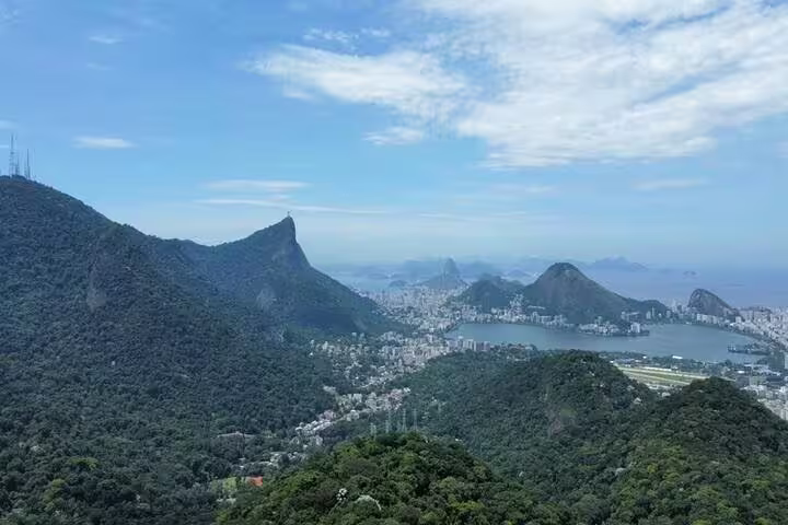 Aerial view of Rio de Janeiro's lush Tijuca Rainforest, with iconic Sugarloaf Mountain and cityscape in the distance.