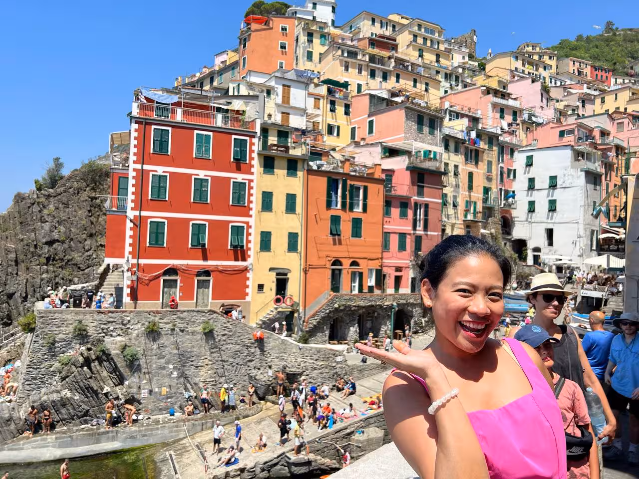 Smiling tourist posing in front of vibrant cliffside buildings at Riomaggiore, Cinque Terre on a sunny day.