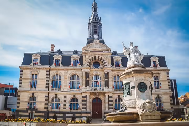 Historic French town hall with clock tower, ornate facade, French flags, and classical fountain statue in foreground