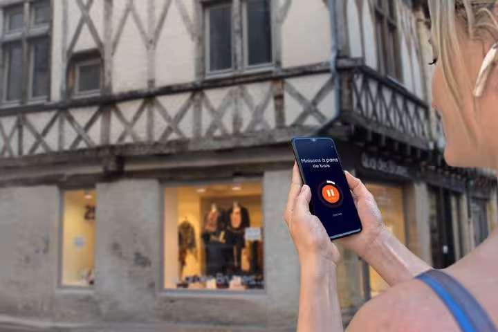 Woman holding smartphone with GPS audio guide app in front of historic half-timbered building in Roanne